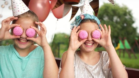 Crazy cheerful girls in birthday hats smiling, having fun and looking through two pink cookies on their eyes. Sweets. Outdoor portrait, kids party, happy birthday