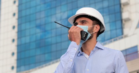 An engineer in a white protective helmet and a face mask speaks on a walkie-talkie in front of a building with blue windows.