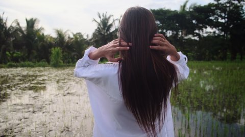 Young woman in white shirt is touching her long hair, walking by a narrow path of green rice paddy fields. Exotic vacation in Bali, Indonesia. Close up, slow motion, back view