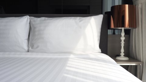 Sunlight through a large window shines on bed with clean white sheets. Lamp on nightstand with copper shade. Modern luxury furnishings in hotel or condo. PAN LEFT.