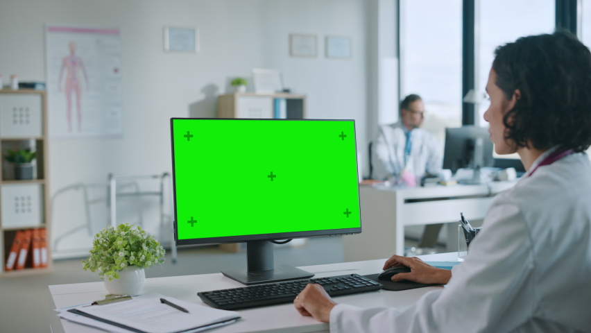 Female Medical Doctor is Working on a Computer with Green Screen Mock Up Display in a Health Clinic. Assistant in White Lab Coat is Reading Medical History Behind a Desk in Hospital Office. | Shutterstock HD Video #1059073805