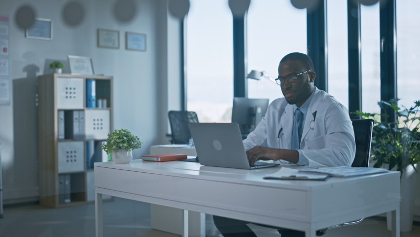 Calm African American Family Medical Doctor in Glasses is Working on a Laptop Computer in a Health Clinic. Physician in White Lab Coat is Browsing Medical History Behind a Desk in Hospital Office. | Shutterstock HD Video #1059073853