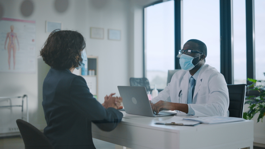 Family Doctor in Protective Mask Reading Medical History of Young Female Patient and Speaking with Her During Consultation in a Health Clinic. Physician in Lab in Front of Computer in Hospital Office. | Shutterstock HD Video #1059073865