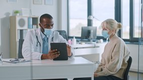 African American Doctor in Protective Mask is Reading Medical History of Senior Female Patient During Consultation in a Health Clinic. Physician Using Tablet Computer in Hospital Office. - Powered by Shutterstock - Get 15% off with code: PIKWIZARD15