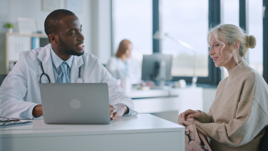 Family Doctor is Reading Medical History of Senior Female Patient and Speaking with Her During Consultation in a Health Clinic. Physician in Lab Coat Sitting Behind a Laptop in Hospital Office.