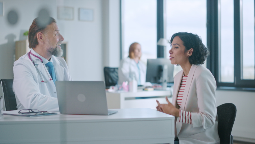 Family Doctor is Delivering Great News About Female Patient's Medical Results During Consultation in a Health Clinic. Physician in White Lab Coat Sitting Behind a Computer in Hospital Office. | Shutterstock HD Video #1059073955
