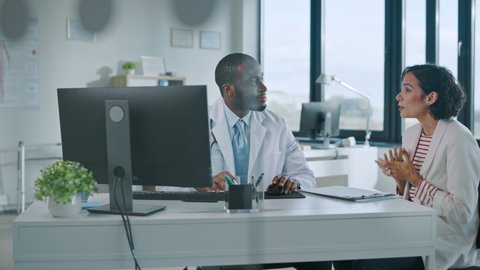 Family Doctor is Delivering Great News About Female Patient's Medical Results During Consultation in a Health Clinic. Physician in White Lab Coat Sitting Behind a Computer in Hospital Office.