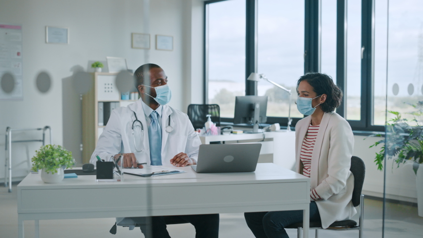 Family Doctor in Protective Mask is Reading Medical History of Female Patient and Speaking with Her During Consultation in a Health Clinic. Physician in Lab in Front of Computer in Hospital Office. | Shutterstock HD Video #1059073985