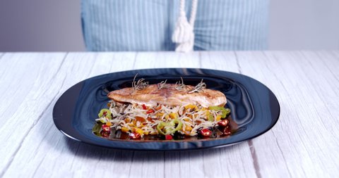 Woman's hand pouring hot spices on a plate of grilled salmon with a base of vegetables and rice.