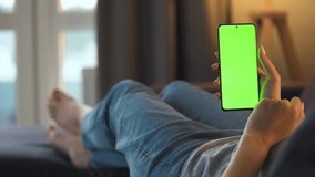 Woman at home lying on a sofa and using smartphone with green mock-up screen in vertical mode. Girl browsing Internet, watching content, videos, blogs. POV.