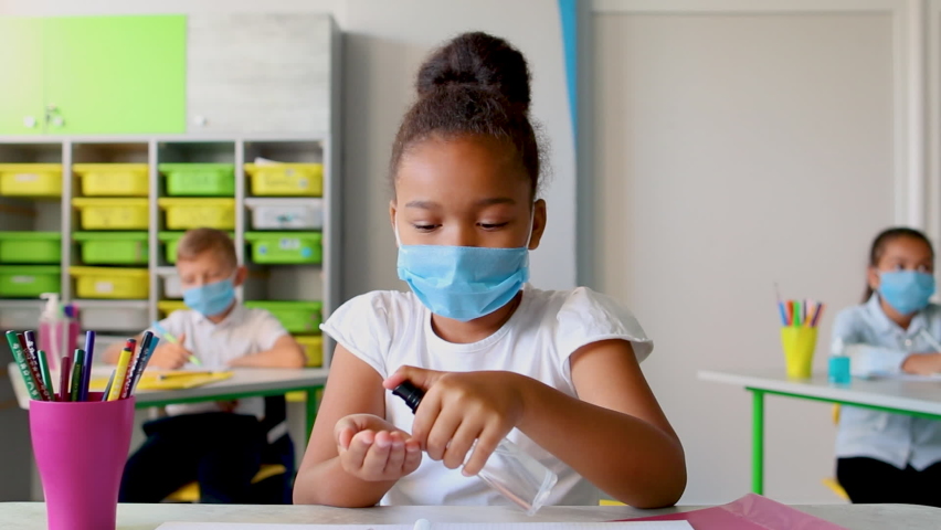 Little black girl with face mask in class puts hand sanitizer on her hands. COVID-19 pandemic. Children back to school after coronavirus lockdown. | Shutterstock HD Video #1059075104