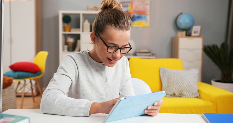 Pretty Caucasian teen little girl in glasses sitting at the table in her cozy room and tapping or scrolling on the tablet device.
