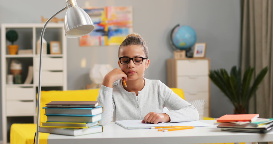 Portrait shot of the cute Caucasian teen girl in glasses and with fair hair sitting at the table in her room at home and doing homework.