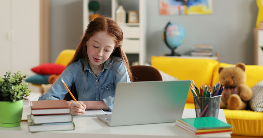 Caucasian schoolgirl with red long hair writing something while doing homework at the table with laptop computer in the cozy room.