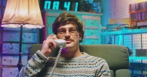 Caucasian funny young man nerd in glasses and with mustache sitting in retro style home and talking on wired vintage telephone of 80's. Male laughing and speak in phone call. Goofy guy of 90's.
