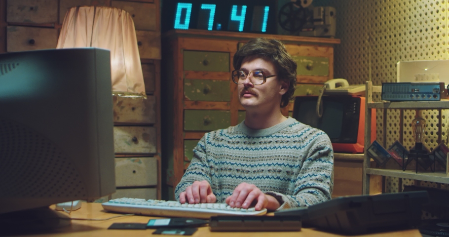 Caucasian male nerd in glasses with mustache sitting at desk in retro room and working on computer. Man programist typing on keyboard and chatting. Vintage style of 90's. Gamer from 80's. | Shutterstock HD Video #1059076091