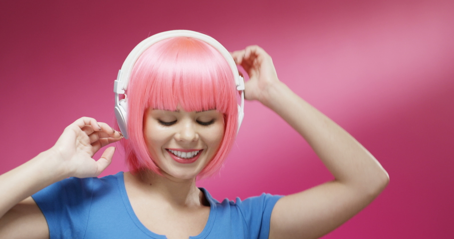 Close up of attractive cheerful young smiled woman in pink short wig and headphones dancing joyfully. Portrait of beautiful hapy gir listening to music and laughing on bright background wall.