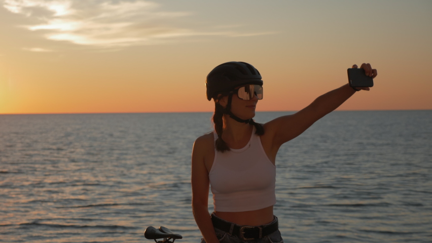 Portrait of young happy woman in helmet glasses and stylish outfit is taking selfies on phone at sunset on sea coastline. Smiling female cyclist takes selfie standing on promenade of sea at sunrise. Royalty-Free Stock Footage #1059078197