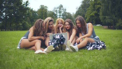 Happy young cheerleaders sitting on grass in park and checking competition results on laptop celebrating victory. Excited cheerleading team happy about winning