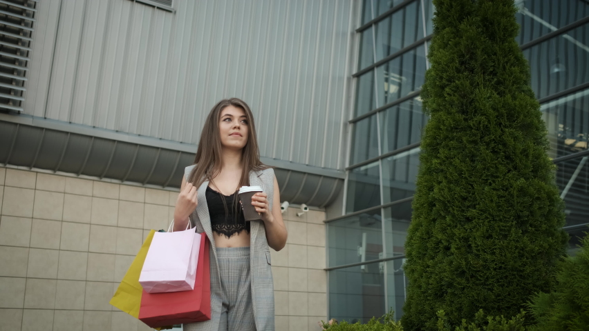 A woman in a stylish gray suit and open neckline returns from shopping and drinks coffee and tea. Gift packages. Modern buildings