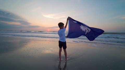Boy standing on Myrtle Beach at sunrise waving South Carolina Flag. . High quality FullHD footage. Boy flying SC flag in morning light on the beach.
