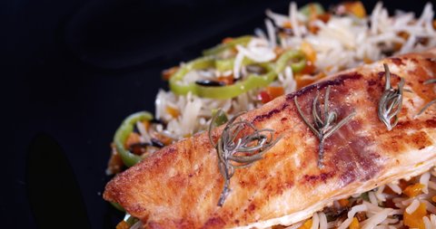 Close-up view of grilled salmon fillet decorated with rosemary and a base of rice and vegetables.