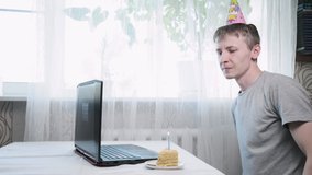 young caucasian man in funny cone hat sitting at table with laptop computer, blowing lighted candle on birthday cake, making wish. Celebrating at home during covid-19 quarantine. Online communication
