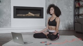 Beautiful young multiracial woman does yoga and strength training exercises on a mat in her living room. She follows an online exercise course video on her laptop.