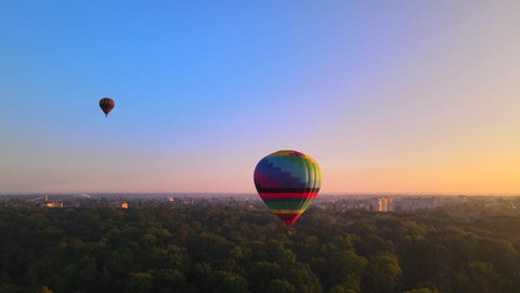 Aerial drone view of colorful hot air balloon flying over green park and river in small european city at summer sunrise, Kiev region, Ukraine