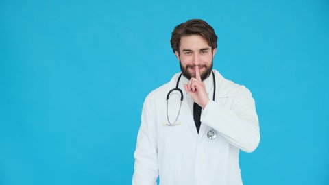 Portrait of serious doctor in professional medical white coat holding finger on lips over blue background. Gesture of shhh, secret, silence. Body language. Doc man