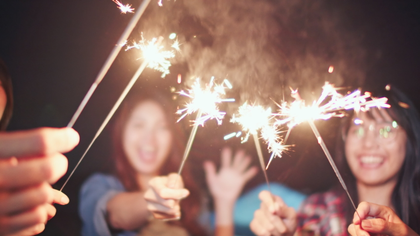 Group of young Asian college student friends lit light sparkler, sing and dance together at beach camping tent. Party people, love friendship relationship, or outdoor camping activity concept