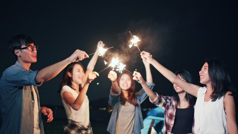 Group of young Asian college student friends lit light sparkler, sing and dance together at beach camping tent. Party people, love friendship relationship, or outdoor camping activity concept