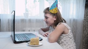 Little caucasian girl in cone hat sitting at table with laptop computer, blowing lighted candle on birthday cake, chats with relatives via video call. Celebrating at home during covid-19 quarantine