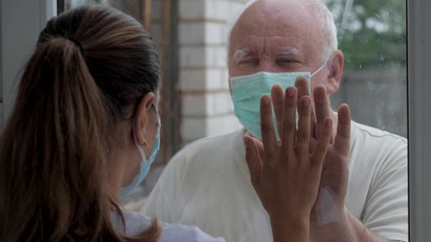 Adult old man in protective medical mask and illness woman touch hands through glass window that separates them. Quarantine for coronavirus pandemic. Hope hand and support for recovery from covid-19