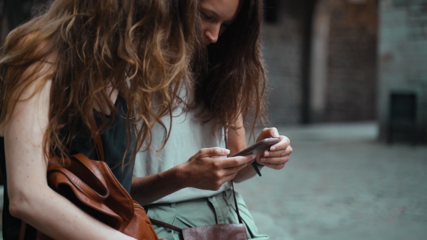 Two friends sit on the edge of a fountain in a beautiful square. Two young long-haired women in the stone medieval town. Two tourists look at the phone screen. Girl in black dress.Girl in grey t-shirt