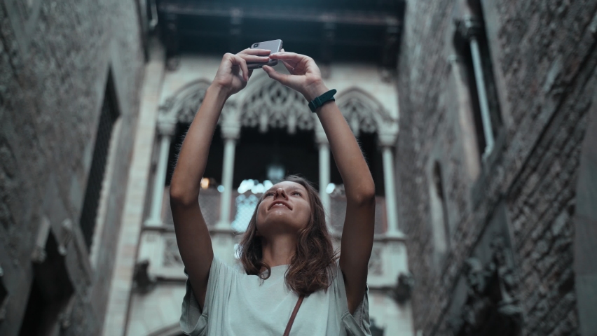 Beautiful long-haired brunette making photo under the arch. Young attractive woman taking pictures of stunning buildings. Pretty tourist makes selfie in a beautiful city. Girl with mobile phone.