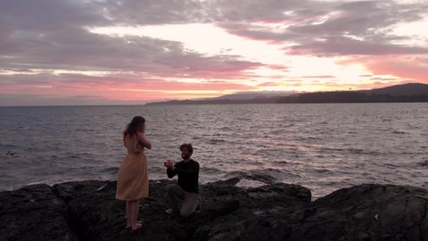 A attractive young man getting on his knee to propose to his girlfriend beside the ocean at sunset. 4k 24fps.