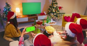 Christmas holidays concept  Asian young friends are watching green chroma key screen TV and using smartphone while sitting on couch in the living room