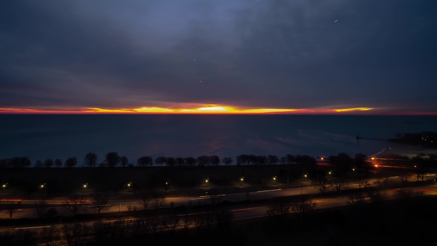 Chicago, IL - March 11th, 2019: The sun rises over the horizon peeping  between the water of Lake Michigan and the cloudy sky above as traffic drives by on Lake Shore Drive as night turns to day.