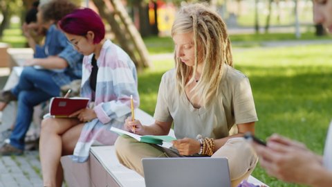 Young female college student sitting with crossed legs on bench in the park and writing in notepad
