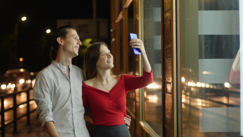 Young happy couple watch store showcase and see expensive thing they wish to buy behing glass frontage during night city walk and take phone picture of it to remember