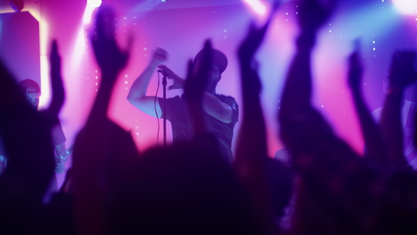 Rock Band with Guitarists and Drummer Performing at a Concert in a Night Club. Front Row Crowd is Partying. Silhouettes of Fans Raise Hands in Front of Bright Colorful Strobing Lights on Stage. Royalty-Free Stock Footage #1059125393
