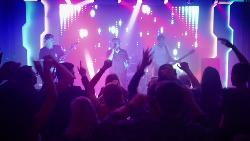 Rock Band with Guitarists and Drummer Performing at a Concert in a Night Club. Front Row Crowd is Partying. Silhouettes of Fans Raise Hands in Front of Bright Colorful Strobing Lights on Stage. | Shutterstock HD Video #1059125435