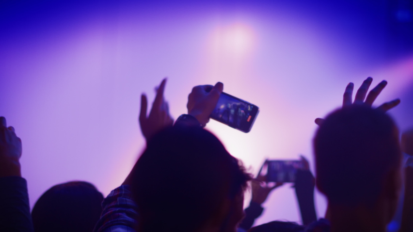 Rock Band with Guitarists and Drummer Performing at a Concert in a Night Club. Front Row Crowd is Record Video on Their Mobile Phones. Party in Front of Bright Colorful Strobing Lights on Stage. Royalty-Free Stock Footage #1059125438