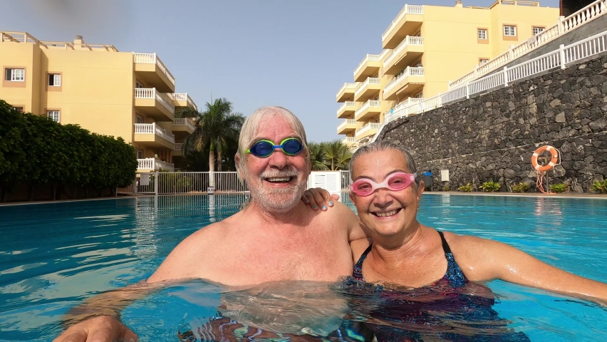couple of seniors enjoying summer and having fun together in the pool swimming and smiling looking at the camera - cheerful happy people in their vacations - portrait of two mature looking underwater Royalty-Free Stock Footage #1059126356