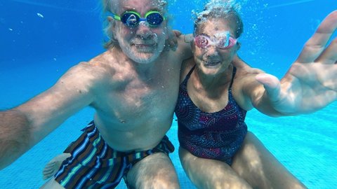couple of seniors enjoying summer and having fun together in the pool swimming and smiling looking at the camera - cheerful happy people in their vacations - portrait of two mature looking underwater