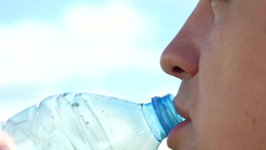 Thirst quenching. Drink clean water from a bottle. A young man of Caucasian appearance drinks water from a transparent plastic bottle against a blue sky with white clouds. Close-up. Royalty-Free Stock Footage #1059127985