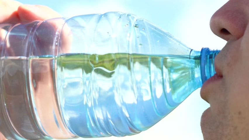 Thirst quenching. Drink clean water from a bottle. A young man of Caucasian appearance drinks water from a transparent plastic bottle against a blue sky with white clouds. Close-up. | Shutterstock HD Video #1059127985