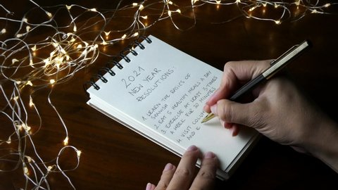 A woman writing a list of things to do or accomplish in 2021, making specific detailed resolutions for the new year, in a notebook in a festive setting with led lights on a dark wooden table.