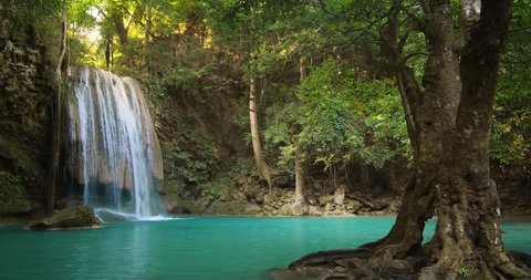 Tropical paradise nature background. Peaceful and tranquil landscape with beautiful waterfall in rain forest of Thailand, emerald pond with turquoise water and old large tree on foreground 